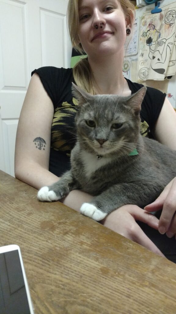 Savanna sitting with a grey cat in her lap at a brown wooden table in front of a fridge with colored illustrations on it.